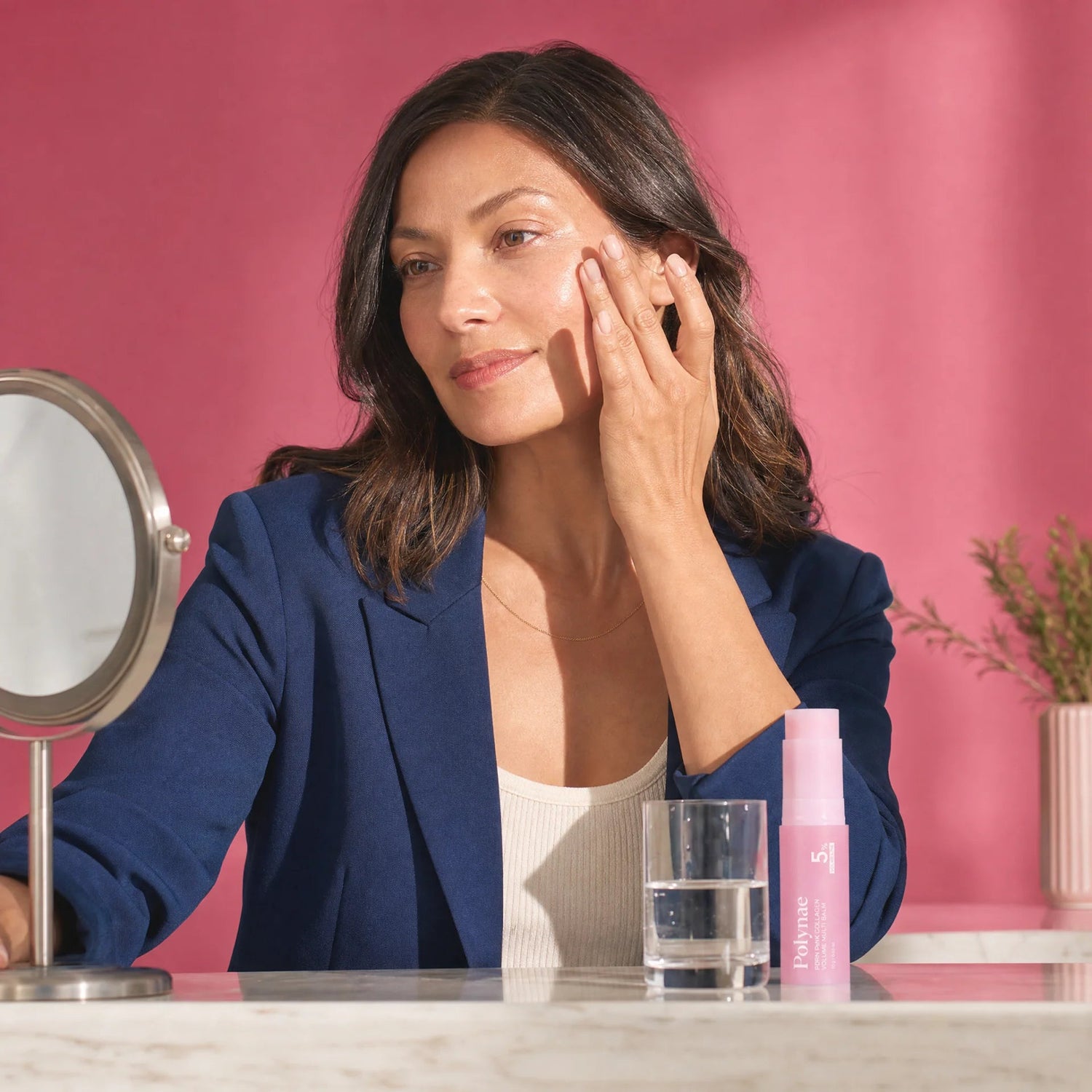 Woman in a blue blazer touching her cheek near a Polynae balm stick and vanity mirror on a marble surface