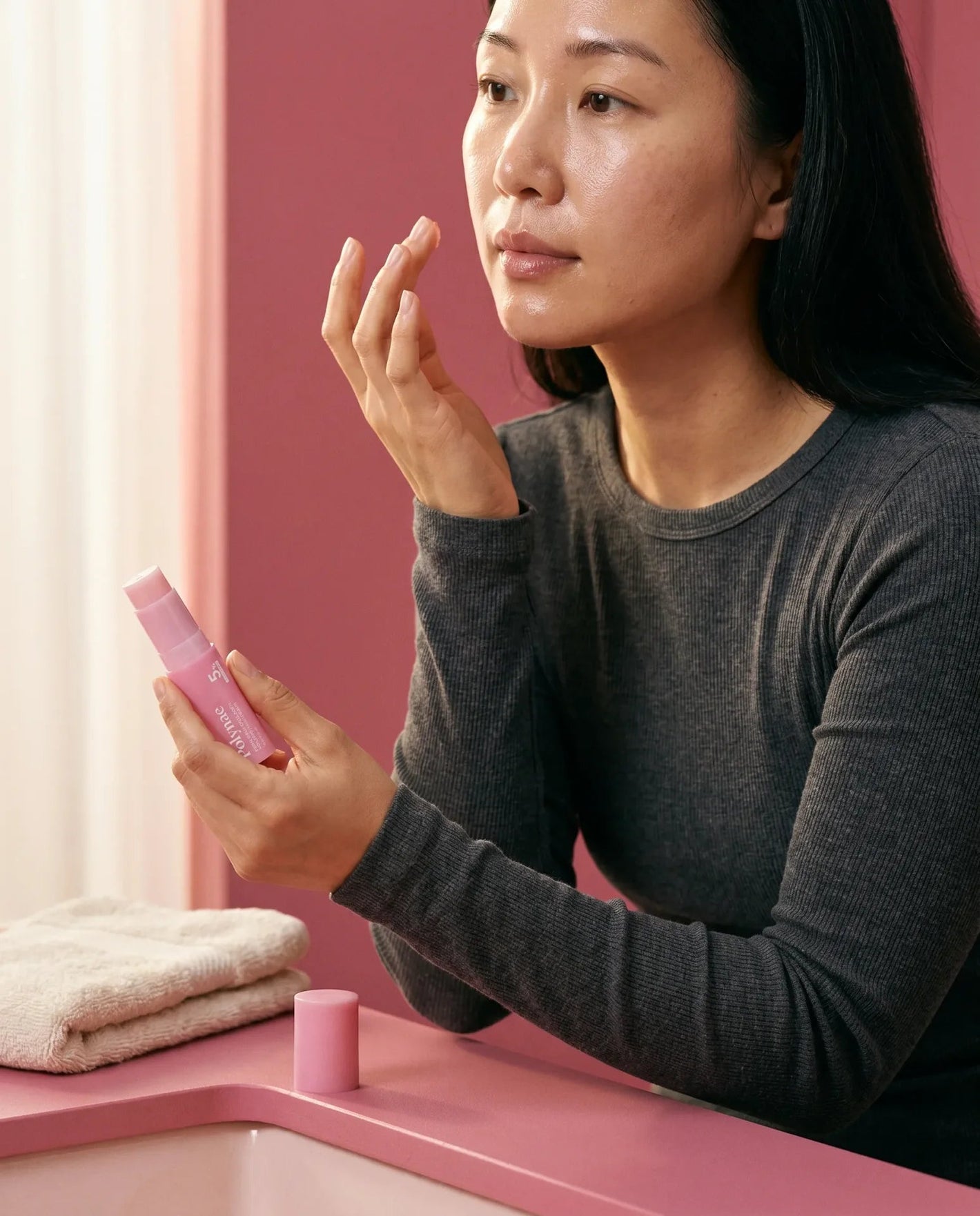 Black woman in rust turtleneck examining a pink Polynae product tube, reflected in a round mirror, pink background