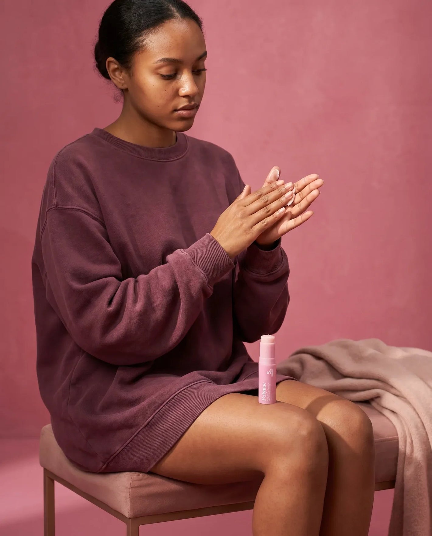 Woman in white shirt standing at pink table with Polynae product and dropper bottles, looking directly at camera
