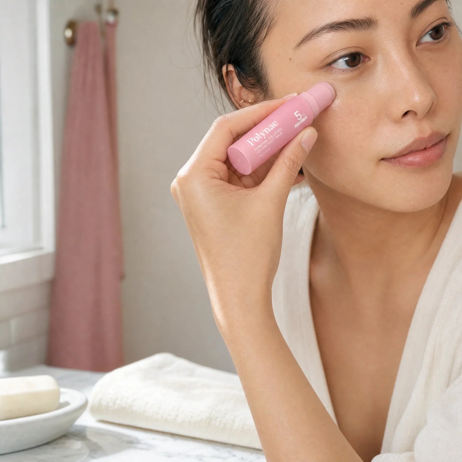 Woman in a white robe applying the Polynae pink balm stick directly to her cheekbone area in a bathroom setting